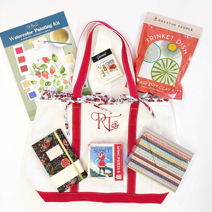 White tote bag with red accents filled with craft items including a watercolor painting kit, air dry clay kit, and other small products.